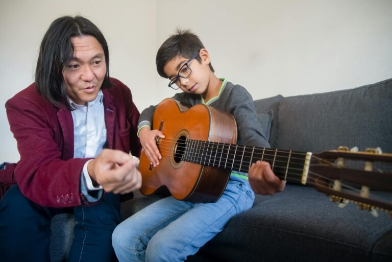 A music teacher guiding a young boy in playing the acoustic guitar indoors.